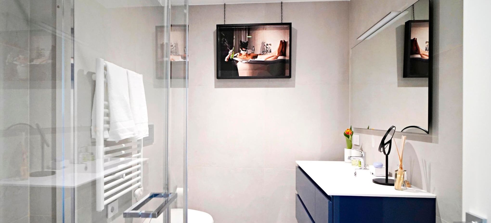 Een moderne badkamer met een glazen douchecabine, een wit wastafeltje met spiegel en een lijstfoto aan de muur.