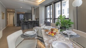Een moderne eetruimte met een groot raam, een keuken op de achtergrond, en een eettafel gedekt met borden, glazen en een mand met fruit.