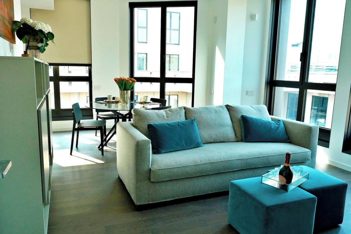 A modern living room with a grey sofa, two blue cushions, a dining table with chairs, and a large window with blinds.