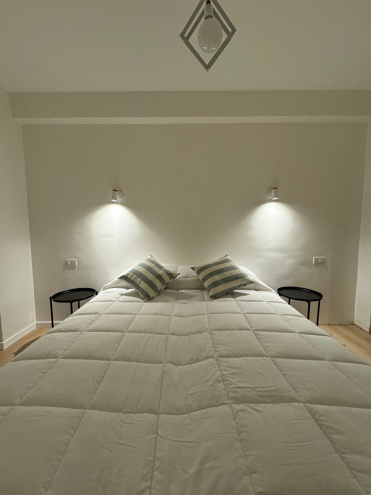 Una cama con edredón blanco acolchado, dos almohadas a rayas y dos mesitas de noche negras.