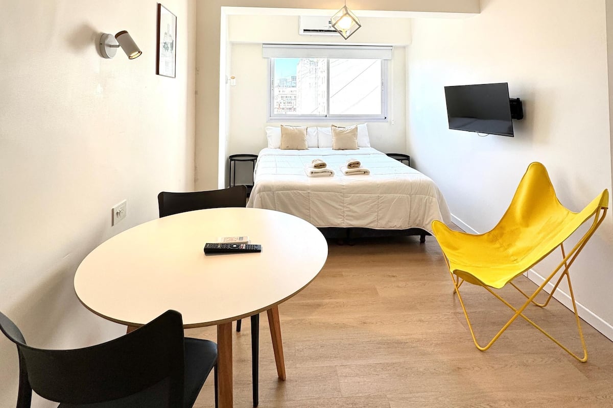 Habitación de hotel pequeña con cama, televisión montada en la pared, mesa de comedor con sillas y una silla de color amarillo brillante.