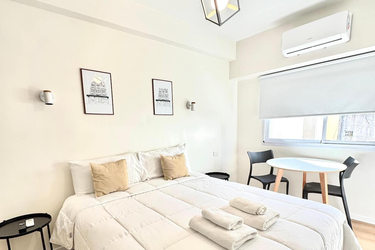 Un dormitorio moderno con una cama grande, una mesita pequeña y una silla. Se ve una ventana con vistas al exterior.