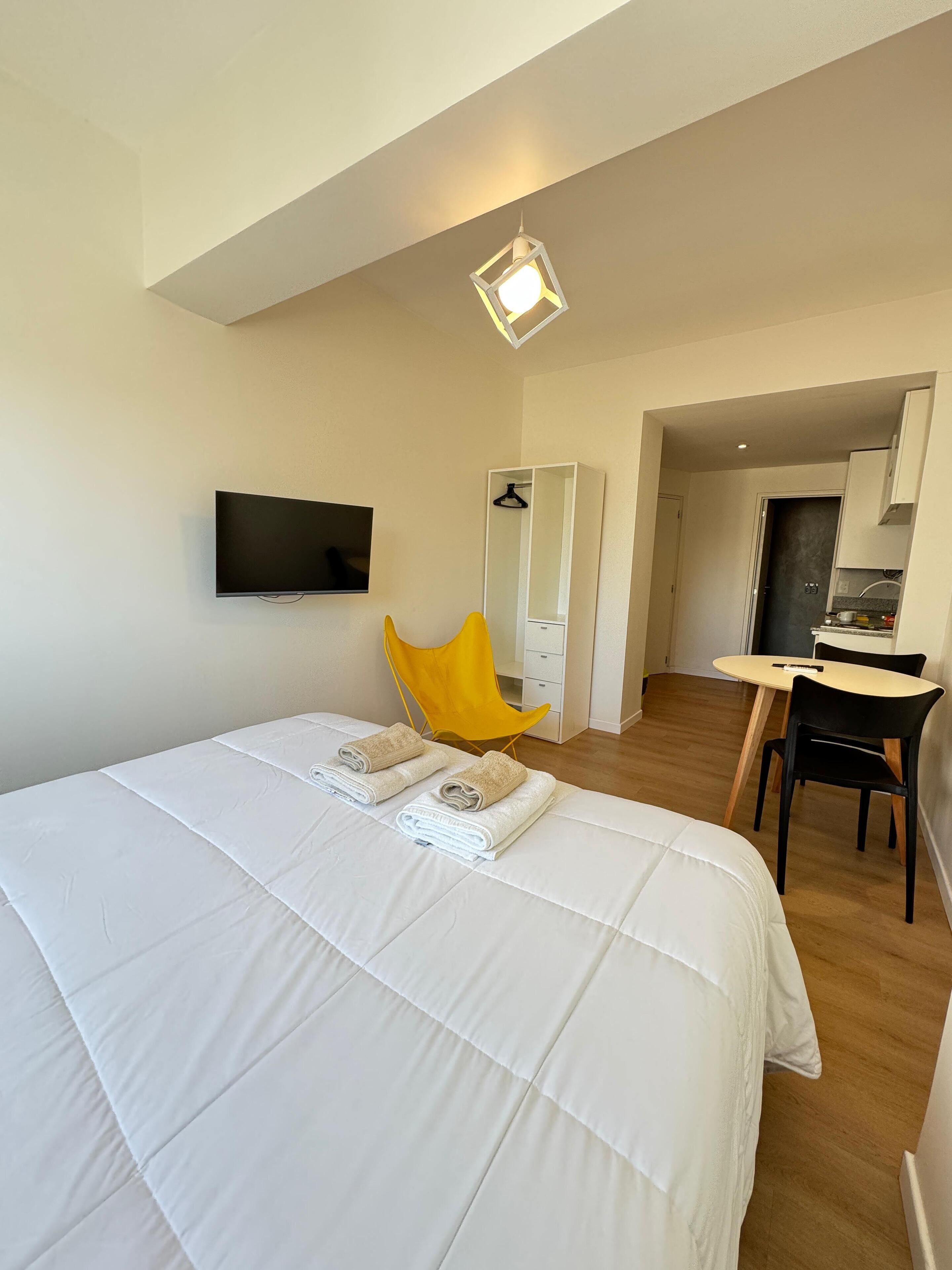 Habitación de hotel con una cama grande, un televisor montado en la pared, una silla amarilla, un escritorio y una kitchenette al fondo.