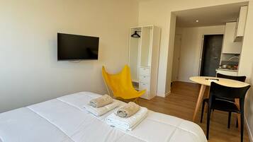 A hotel room with a large bed, a TV mounted on the wall, a yellow chair, a desk, and a kitchenette in the background.