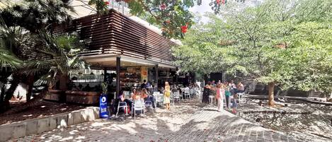 An outdoor café with tables and chairs, surrounded by trees and a brick pathway.