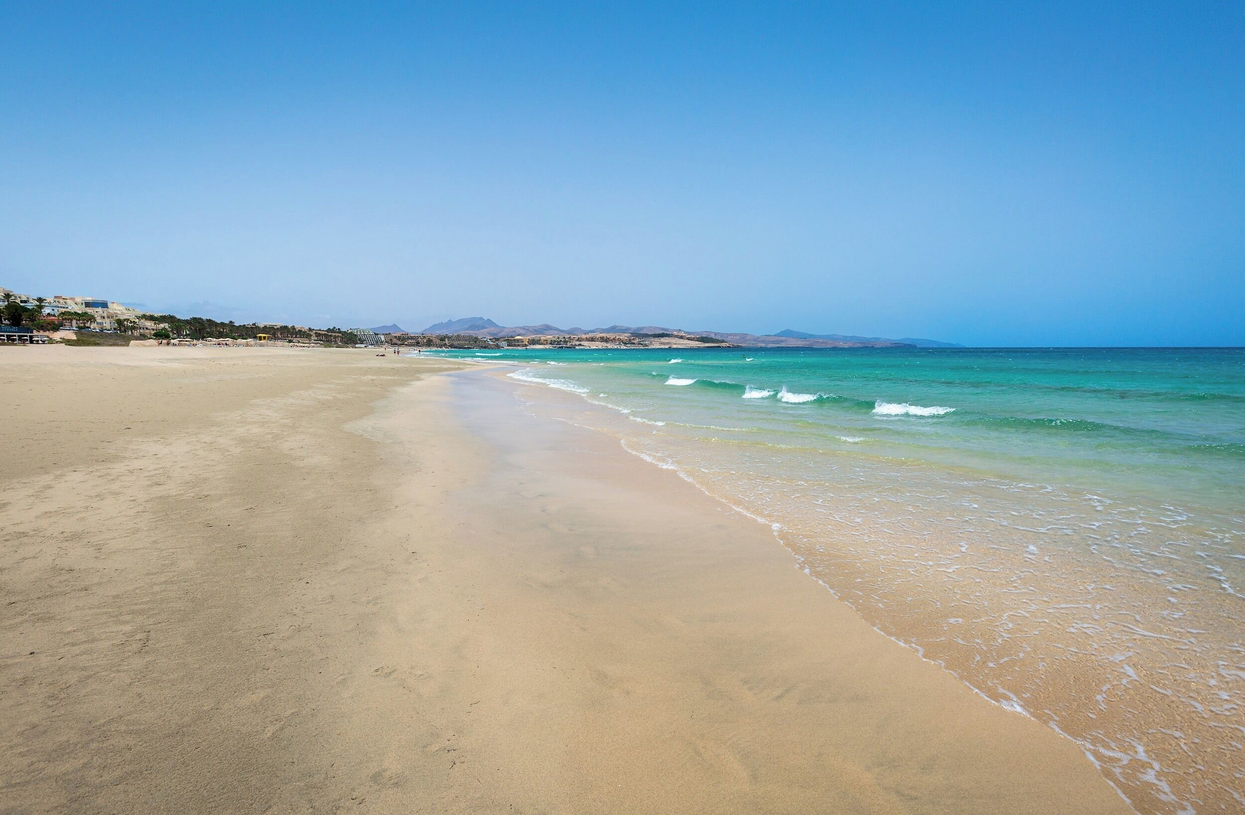 A wide sandy beach with gentle waves, a clear sky, and distant mountains.