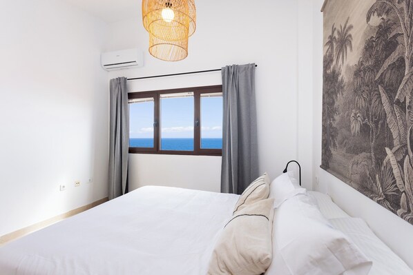 A bedroom with a bed, a window with curtains, a wall-mounted air conditioner, and a tapestry.