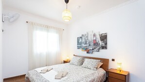 Une chambre à coucher bien aménagée, avec un grand lit, deux tables de chevet avec des lampes, un ventilateur de plafond et une œuvre d’art fixée au mur représentant un paysage urbain.