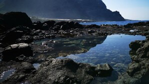 Eine felsige Küste mit klarem Wasser, das die Landschaft spiegelt, ein großer Berg im Hintergrund sowie ein Blick auf einen Strand und Gebäude in der Ferne.