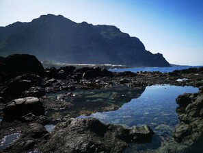 A rocky shoreline with clear water reflecting the landscape, a large mountain in the background, and a distant view of a beach and buildings. - Home2Book Tranquil Spot Buenavista, BBQ & Views (Buenavista del Norte)