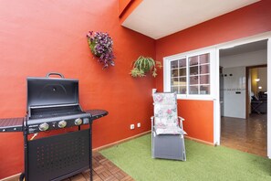 Eine Terrasse mit Grill, einem Stuhl und Kübelpflanzen vor einer roten Wand.
