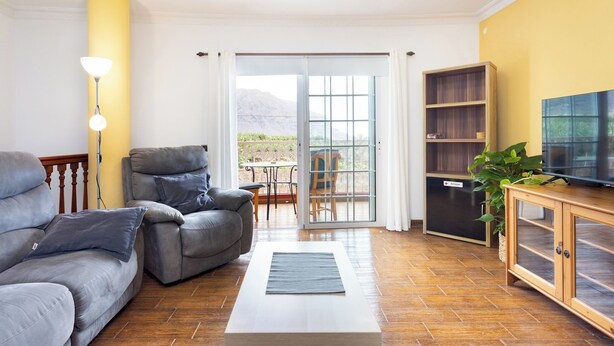 Un salon comprenant un canapé, un fauteuil et un téléviseur posé sur un meuble en bois. Une porte-fenêtre coulissante donne accès à un espace extérieur.