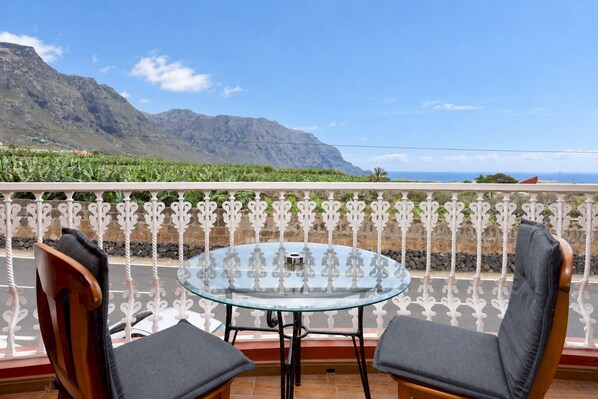 A balcony with a glass table and two chairs, overlooking a mountainous landscape.