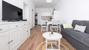 Una sala de estar moderna con televisor de pantalla plana, armarios blancos, un sofá gris y una zona de comedor con mobiliario blanco.