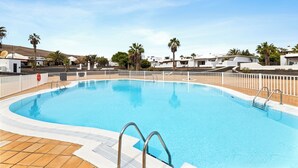 A swimming pool surrounded by white buildings, palm trees, and a clear sky.