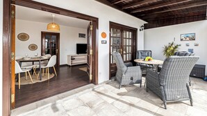 A dining area with wicker chairs and a table, a kitchen with a TV, and a ceiling with exposed beams.
