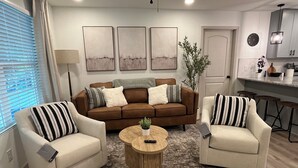 Een woonkamer met een bruine bank, twee fauteuils, een salontafel en een vloerkleed. Er hangen drie abstracte schilderijen aan de muur, en er staat een plant. Ook is er een keuken met barstoelen.