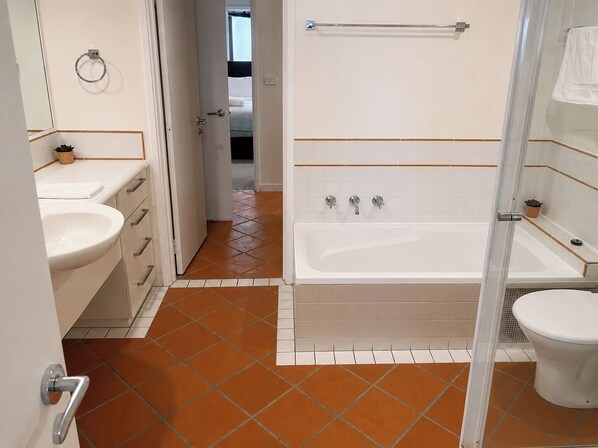 Une salle de bain avec une baignoire, des toilettes et un lavabo, dotée d’un carrelage au sol et d’une porte visible donnant sur une autre pièce.
