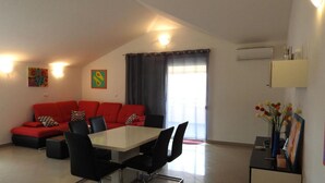 A modern living room with a red sofa, a glass dining table, and black chairs. There are two paintings on the walls, a window with curtains, and an air conditioning unit. - Expansive Ciovo Apartment with Sea-Views, Balcony and Outdoor Dining - Three Bedroom Apartment (Okrug Gornji)