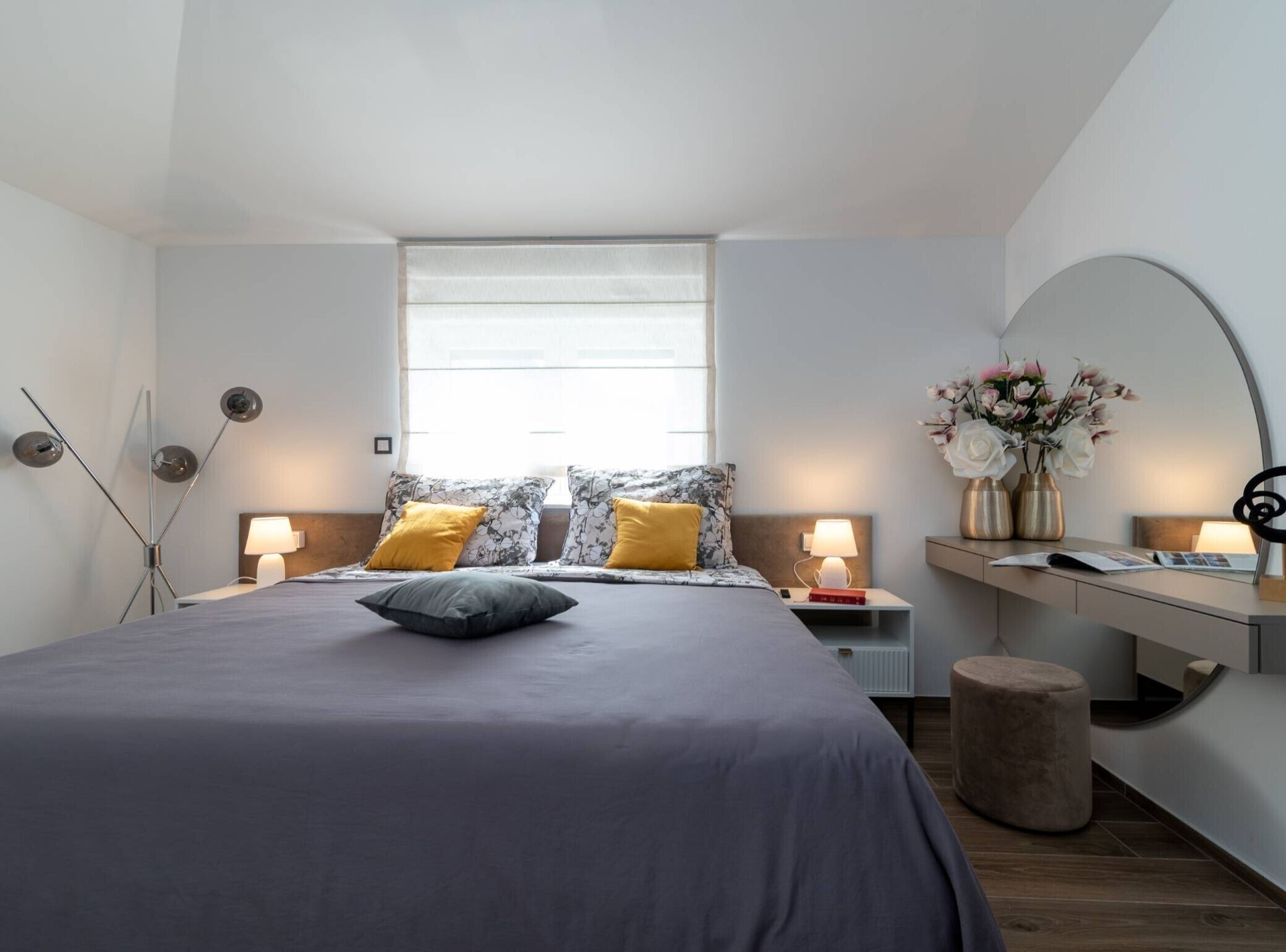 A modern bedroom with a large bed, bedside lamps, a desk with a chair, and a window with curtains.