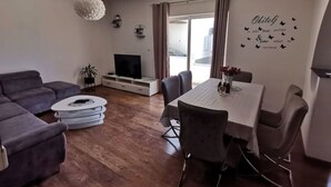 Uma sala de estar moderna com um sofá cinzento, uma mesa de jantar branca com cadeiras, uma TV sobre um suporte branco e uma luminária no teto.