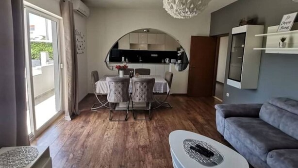 Uma sala de estar moderna com um sofá cinzento, uma área de jantar com mesa e cadeiras, uma cozinha ao fundo e um lustre decorativo grande.