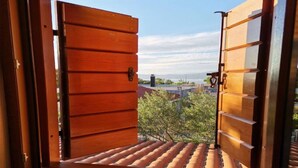 A window with wooden shutters open, revealing a view of a tiled roof, trees, and distant buildings. - Four Bedroom Holiday Home (Skrip)