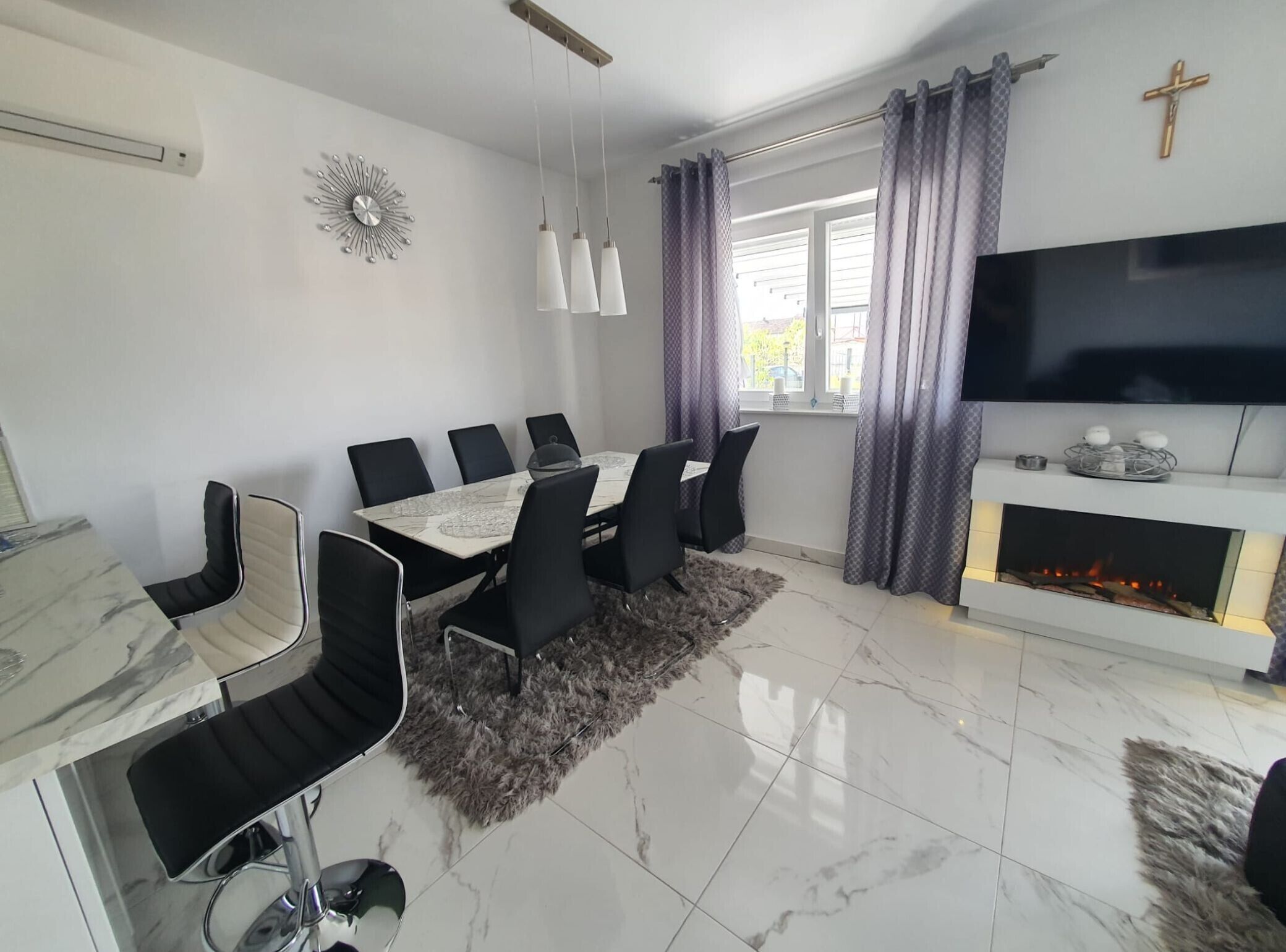 A modern dining room with a marble table, black chairs, a fireplace, and a TV mounted on the wall.