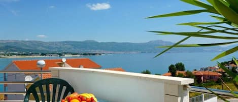 A balcony with a table set for a meal, overlooking a coastal view with buildings and mountains in the distance.