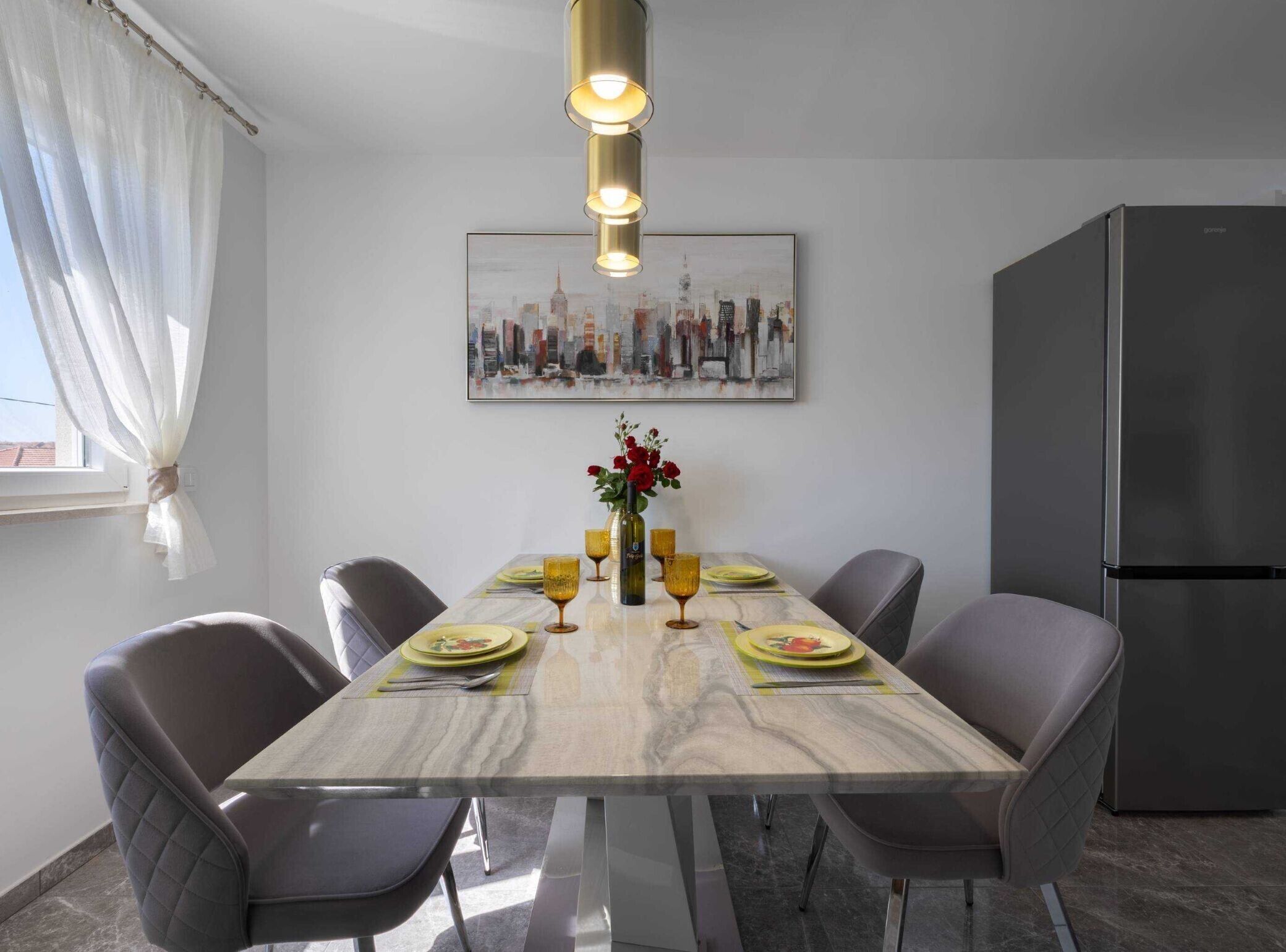 Un espace repas moderne comprenant une table en marbre, six chaises, un lustre, un tableau représentant un paysage urbain et un réfrigérateur.