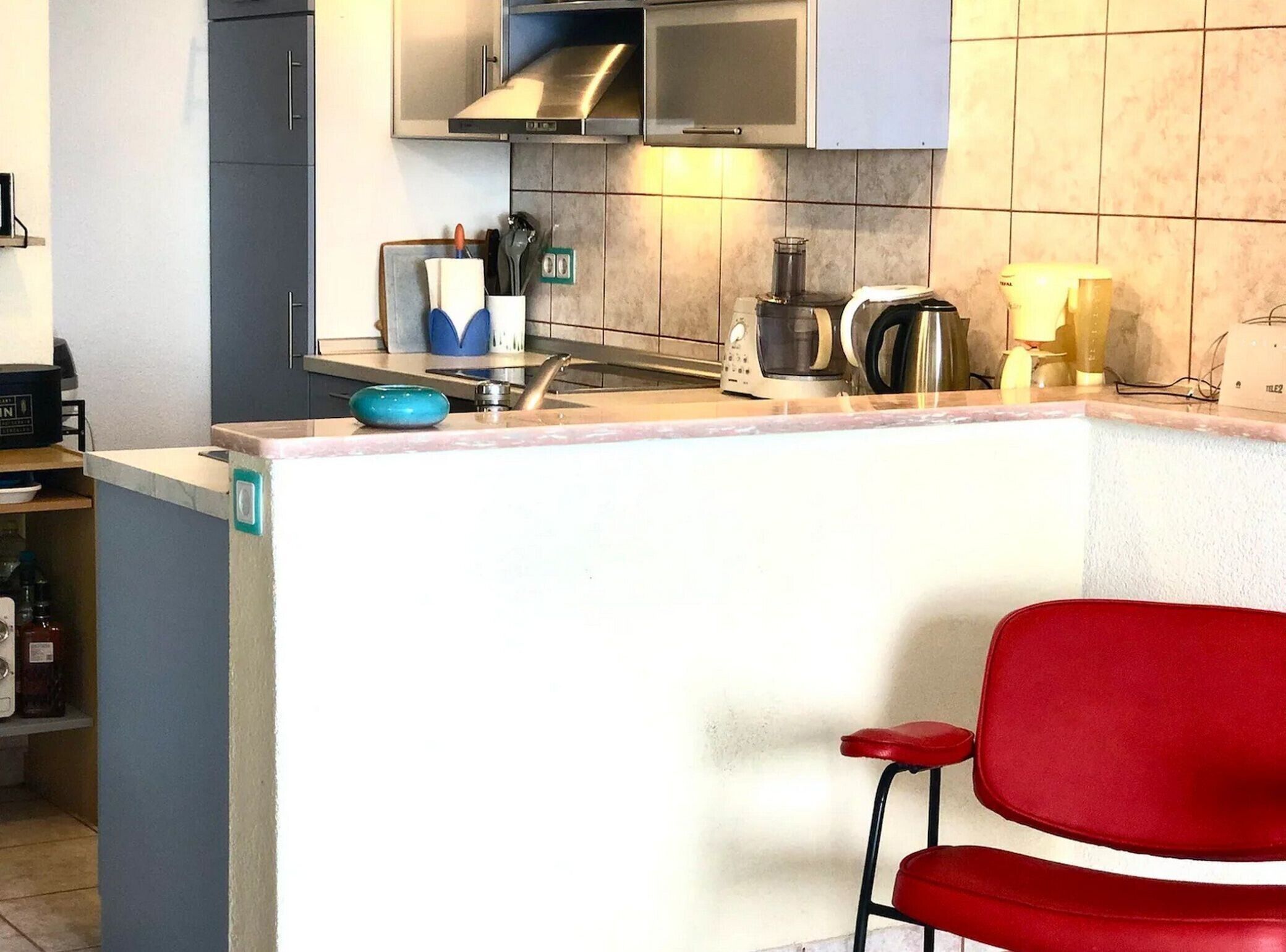 Een keuken met grijze kastjes, een magnetron, een waterkoker en een koffiezetapparaat. Op het aanrecht staan een rode stoel en een blauwe kom.