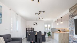 Ett modernt vardagsrum med en grå soffa, ett runt soffbord och en matplats med ett bord och stolar. I bakgrunden syns ett kök med en tegelvägg och hängande lampor.