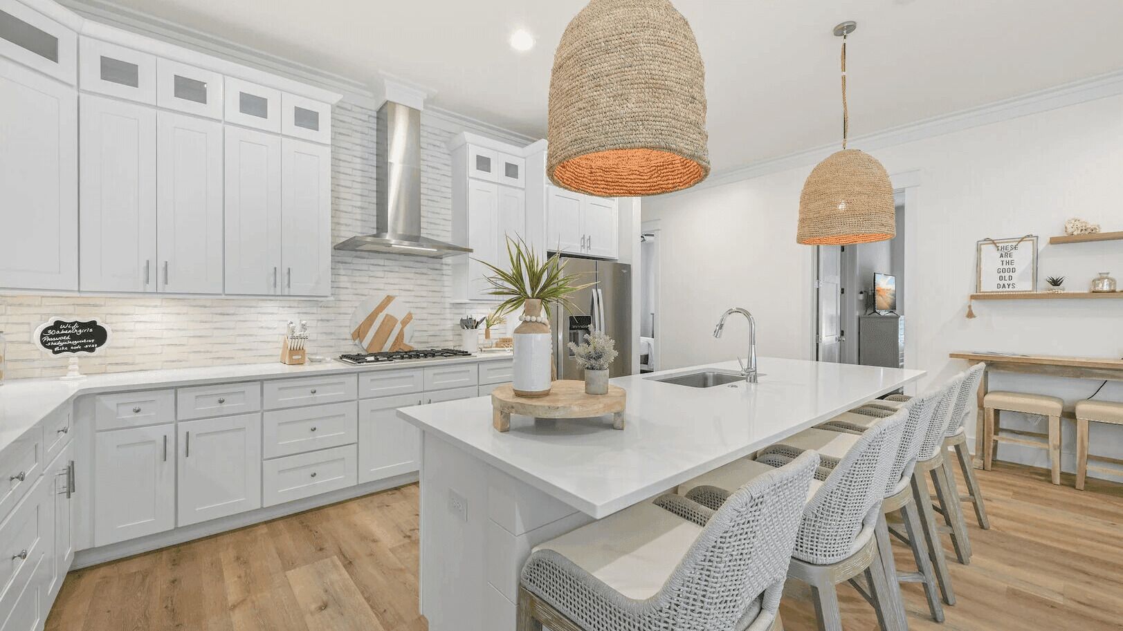 A modern kitchen with white cabinetry, a central island, hanging woven light fixtures, and a wooden bench seating area.