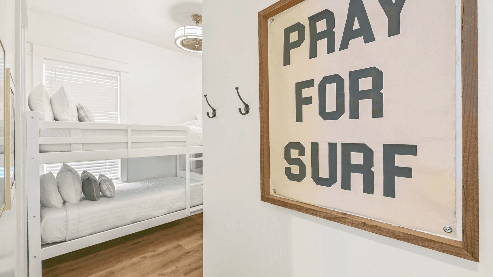 A bunk bed with pillows, a framed sign saying 'PRAY FOR SURF', and a window with blinds.
