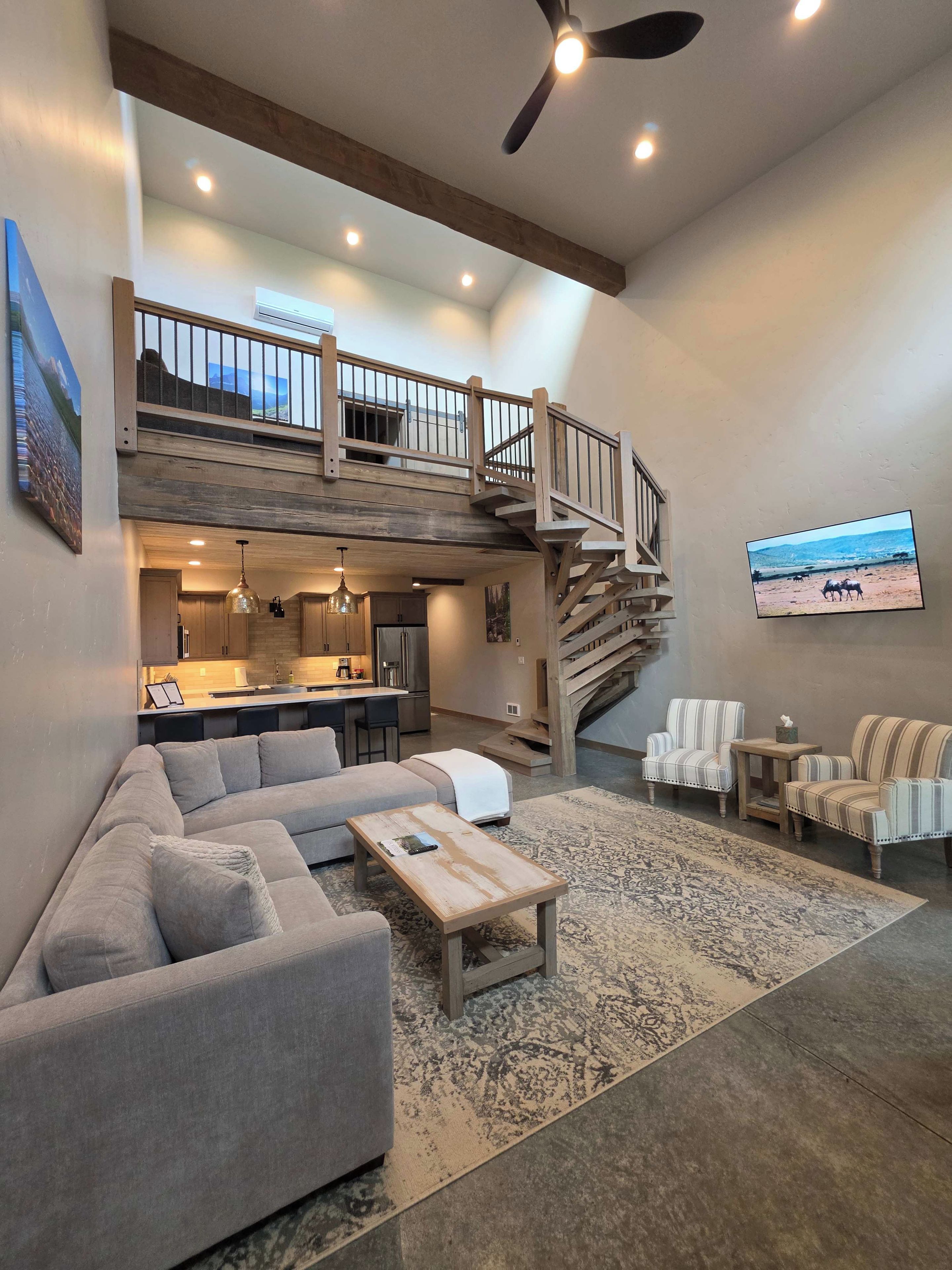 A spacious living area with a high ceiling, a built-in kitchen, a staircase leading to an upper level, and a large flat-screen TV mounted on the wall.