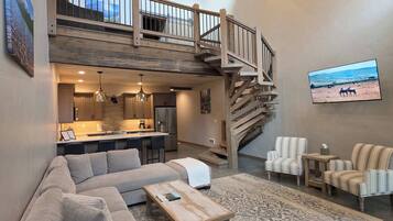 A spacious living area with a high ceiling, a built-in kitchen, a staircase leading to an upper level, and a large flat-screen TV mounted on the wall.