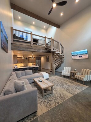 Un'ampia zona giorno con soffitto alto, cucina integrata, una scala che conduce a un livello superiore e un grande televisore a schermo piatto fissato al muro.