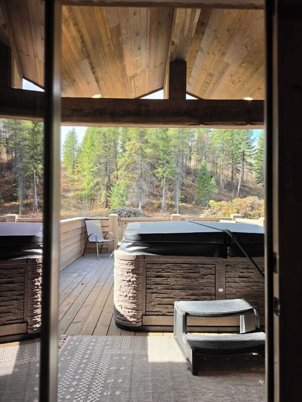 Una terraza de madera con una barbacoa de piedra, mobiliario exterior y vistas a un bosque.