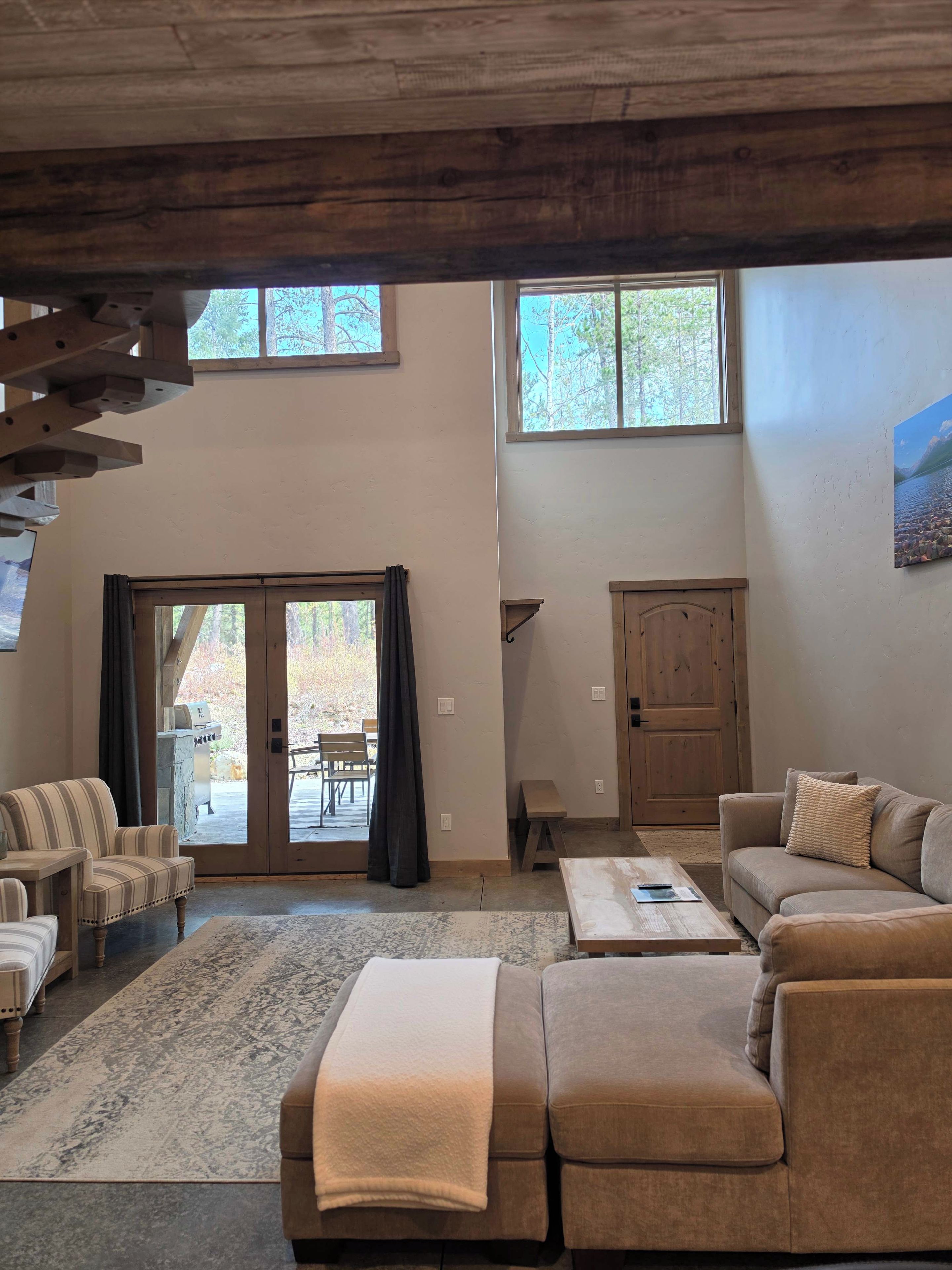 A spacious living room with a large sofa, a coffee table, and a fireplace. There are two windows providing natural light and a view of the outdoors.
