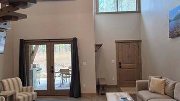 A spacious living room with a large sofa, a coffee table, and a fireplace. There are two windows providing natural light and a view of the outdoors.