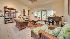 A spacious living room with a piano, wicker furniture, a bookshelf, and a large window. - Lanai + Ocean Views: Secluded Holualoa Retreat! (Holualoa)