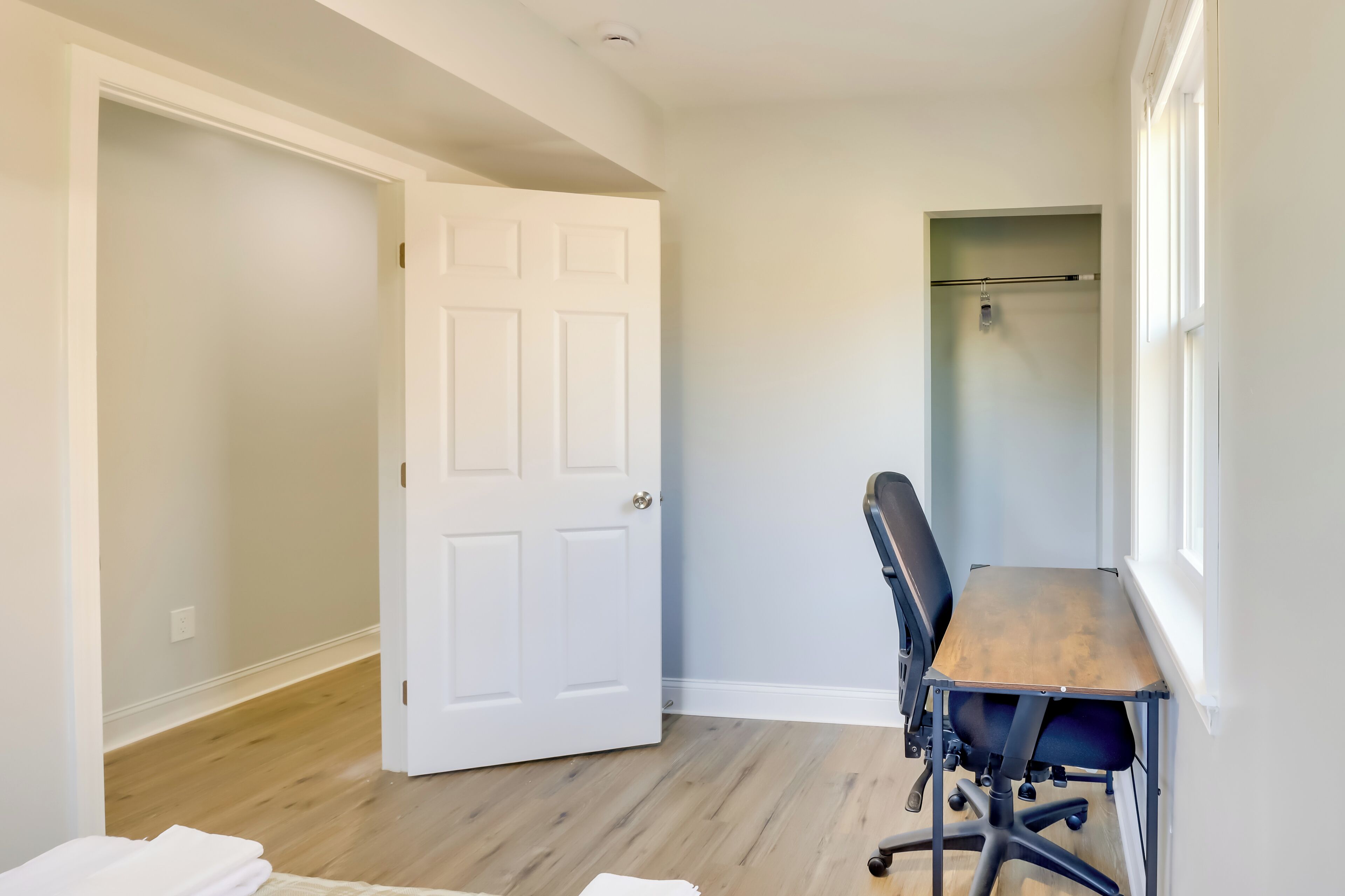 A room with a wooden floor, a white door, a black office chair, and a window with a view.