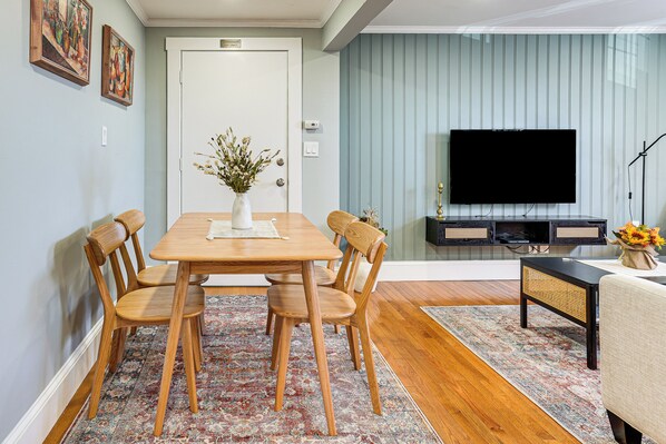 Een eetgedeelte met een houten tafel en stoelen, een vaas met bloemen, een televisie en een bank.