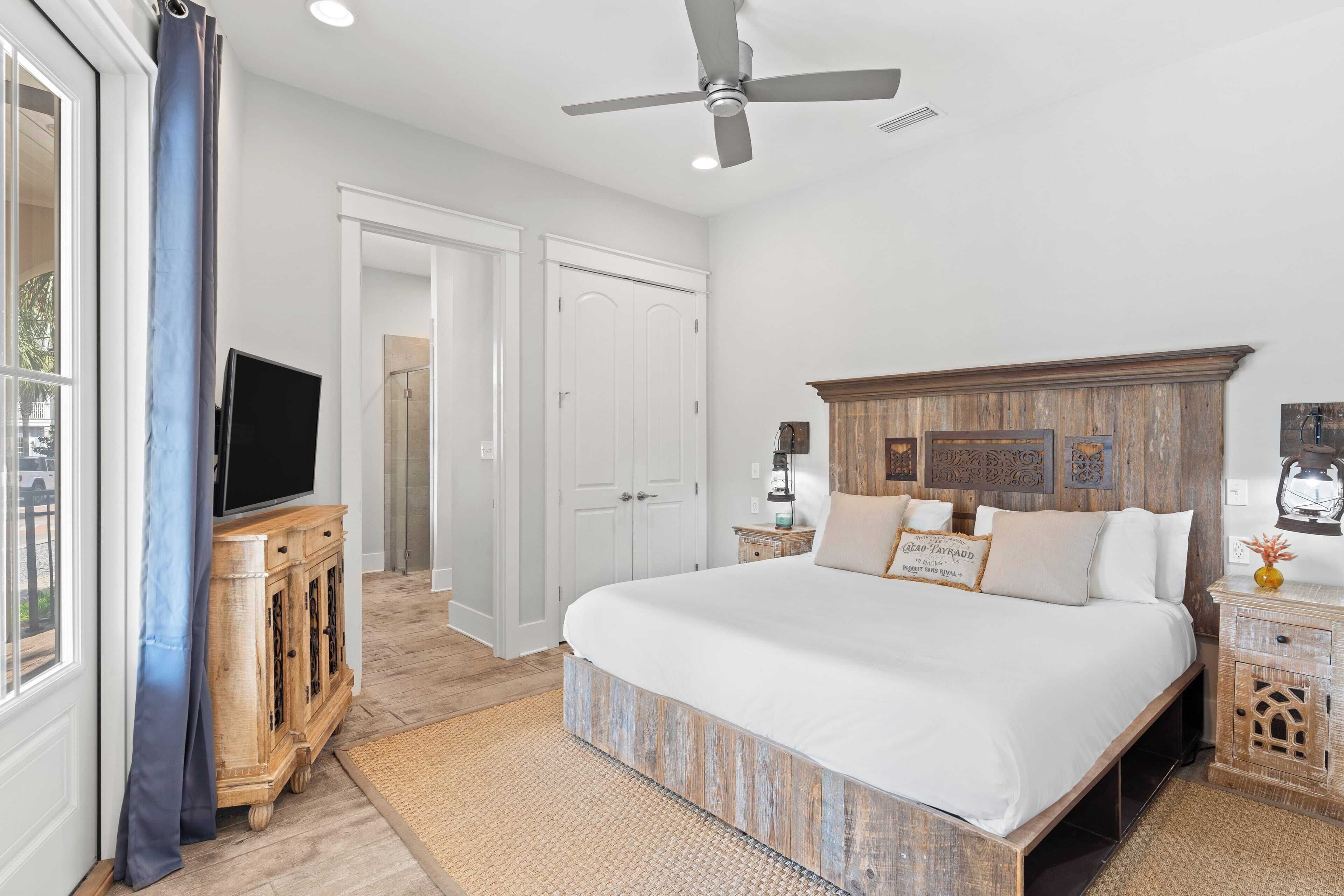A bedroom with a large bed, a ceiling fan, a TV on a wooden cabinet, and a window with curtains.