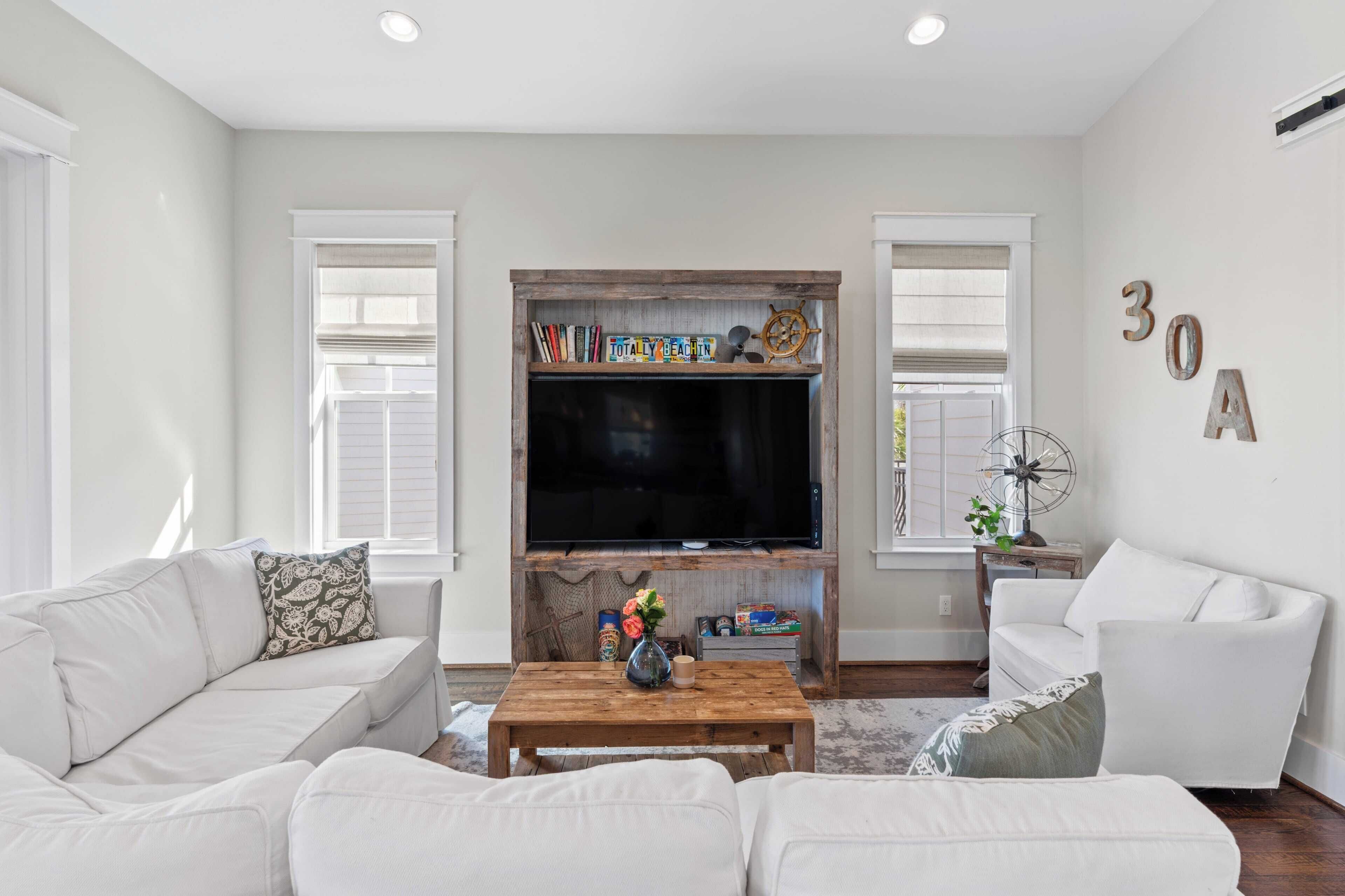 A living room with a large flat-screen TV, white sofas, a wooden coffee table, and a bookshelf with decorative items.