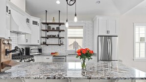 A modern kitchen with white cabinetry, a stainless steel refrigerator, a marble countertop, and a view of a bridge through a window. - Totally Beachin by Avantstay Sleeps 18 Seacrest Beach (Panama City Beach)