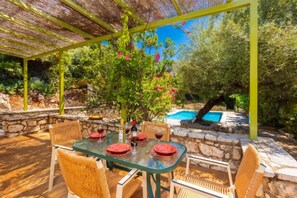 Une terrasse extérieure avec une table dressée pour deux personnes, entourée de chaises en osier, d’un mur de pierre et, au fond, d’une piscine.