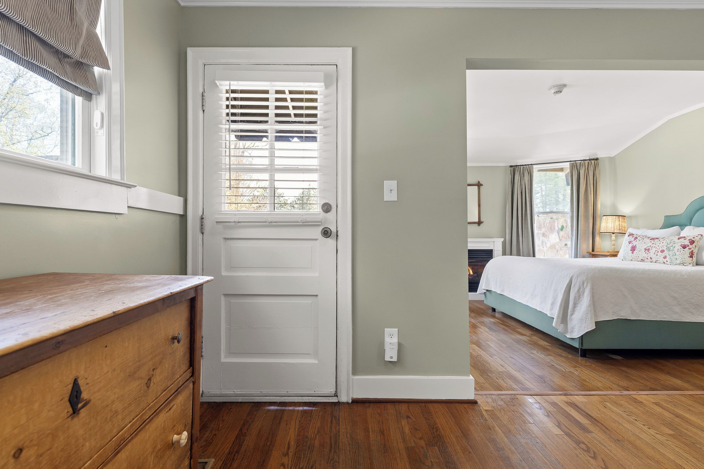 A bedroom with a wooden dresser, a white door with blinds, a bed with a green headboard, and a nightstand with a lamp.