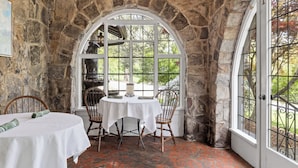 A stone-walled room with a round table and chairs, a large arched window, and a door with a metal gate. - Chanticleer Inn Bed & Breakfast (Lookout Mountain)