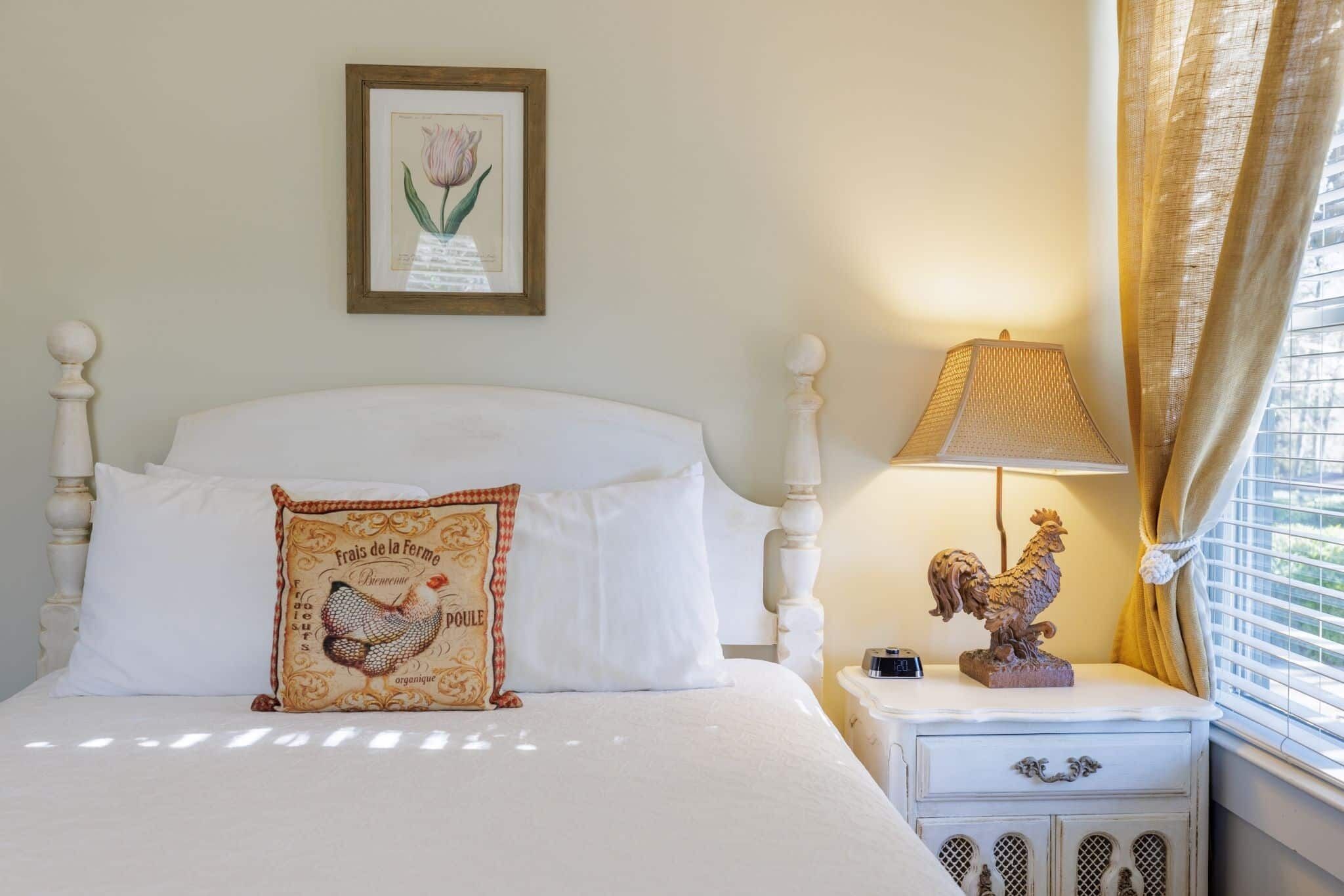 A neatly made bed with white linens and a decorative pillow, a bedside lamp, and a rooster statue on a nightstand.